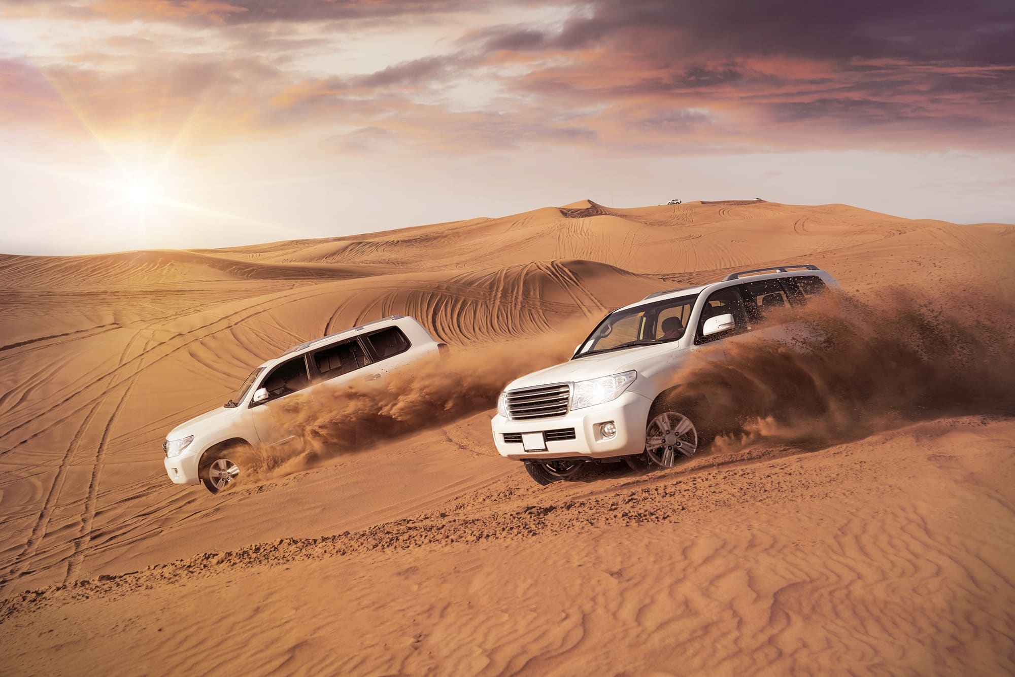 Two four-wheel-drives throwing up plumes of sand as they cross a red dune at sunset, tyre tracks scored across the ridges behind them.