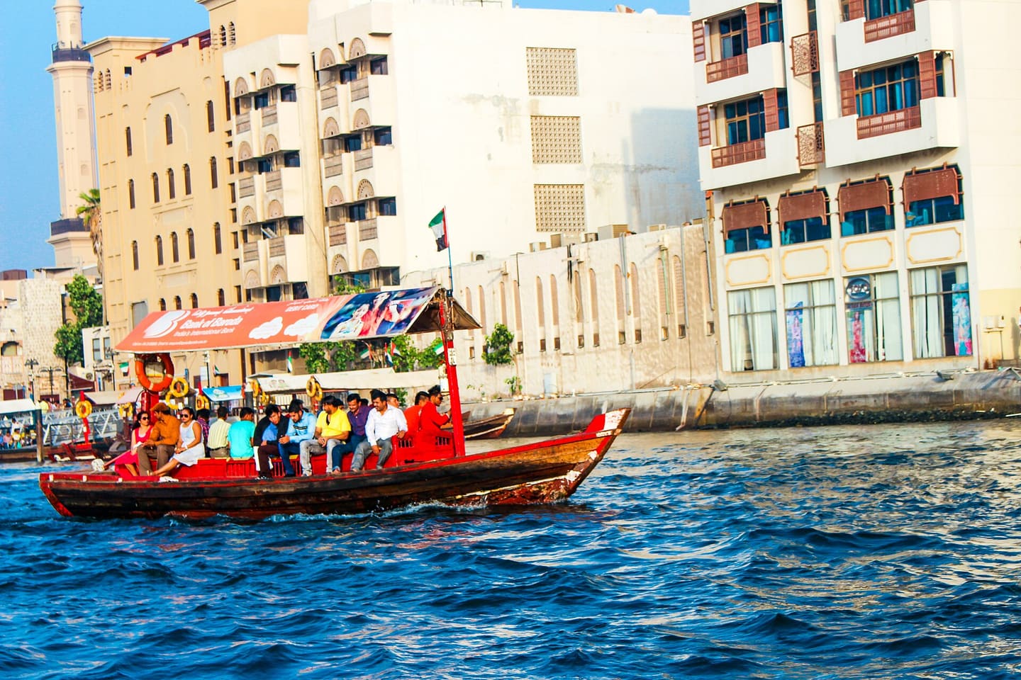 A traditional wooden abra ferry crosses Dubai Creek, passengers seated under its canopy, the Deira waterfront behind.