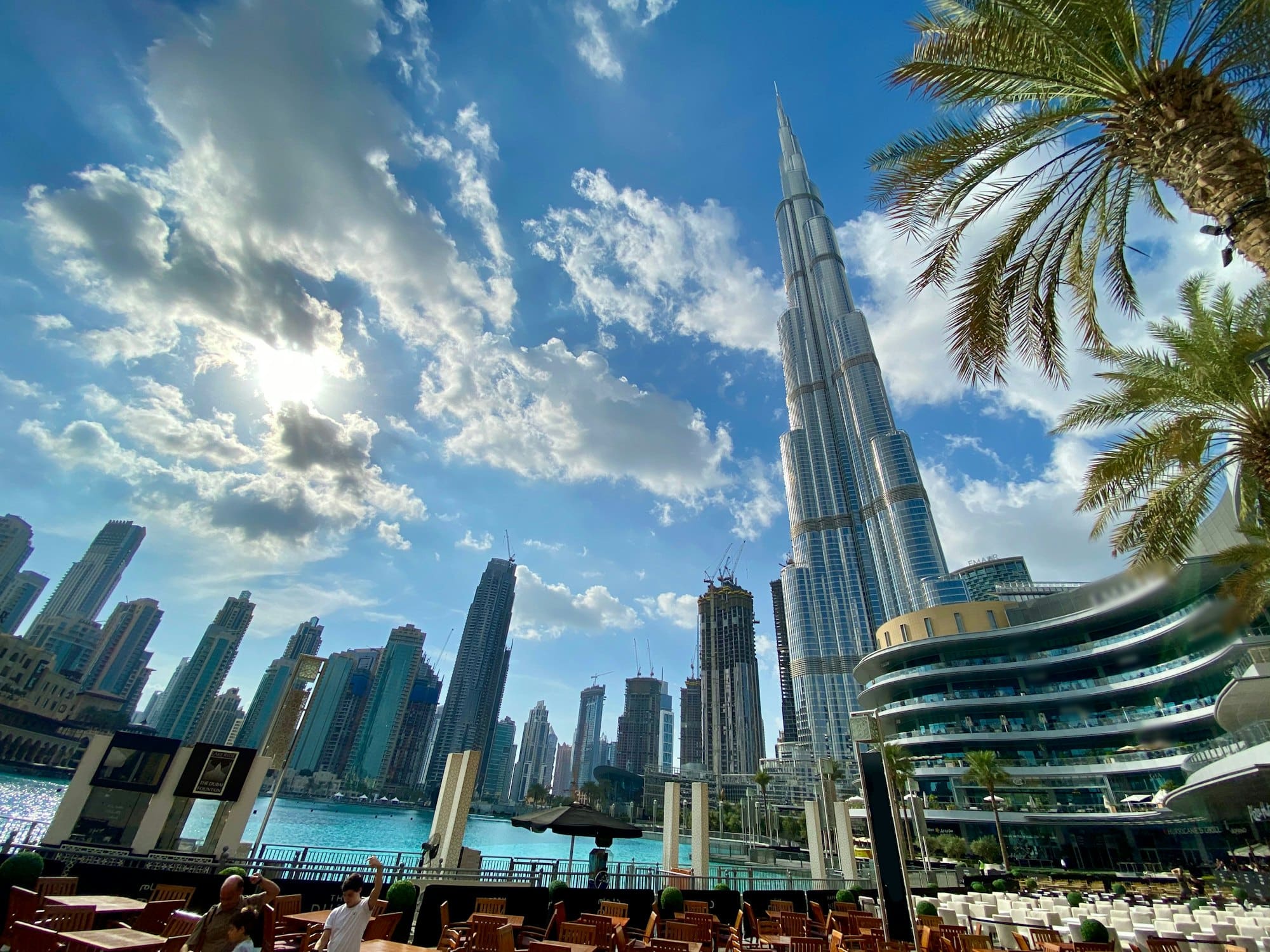 The Burj Khalifa rising above the Dubai Mall lake in afternoon light, palm fronds in the foreground and the fountain lake turquoise below.